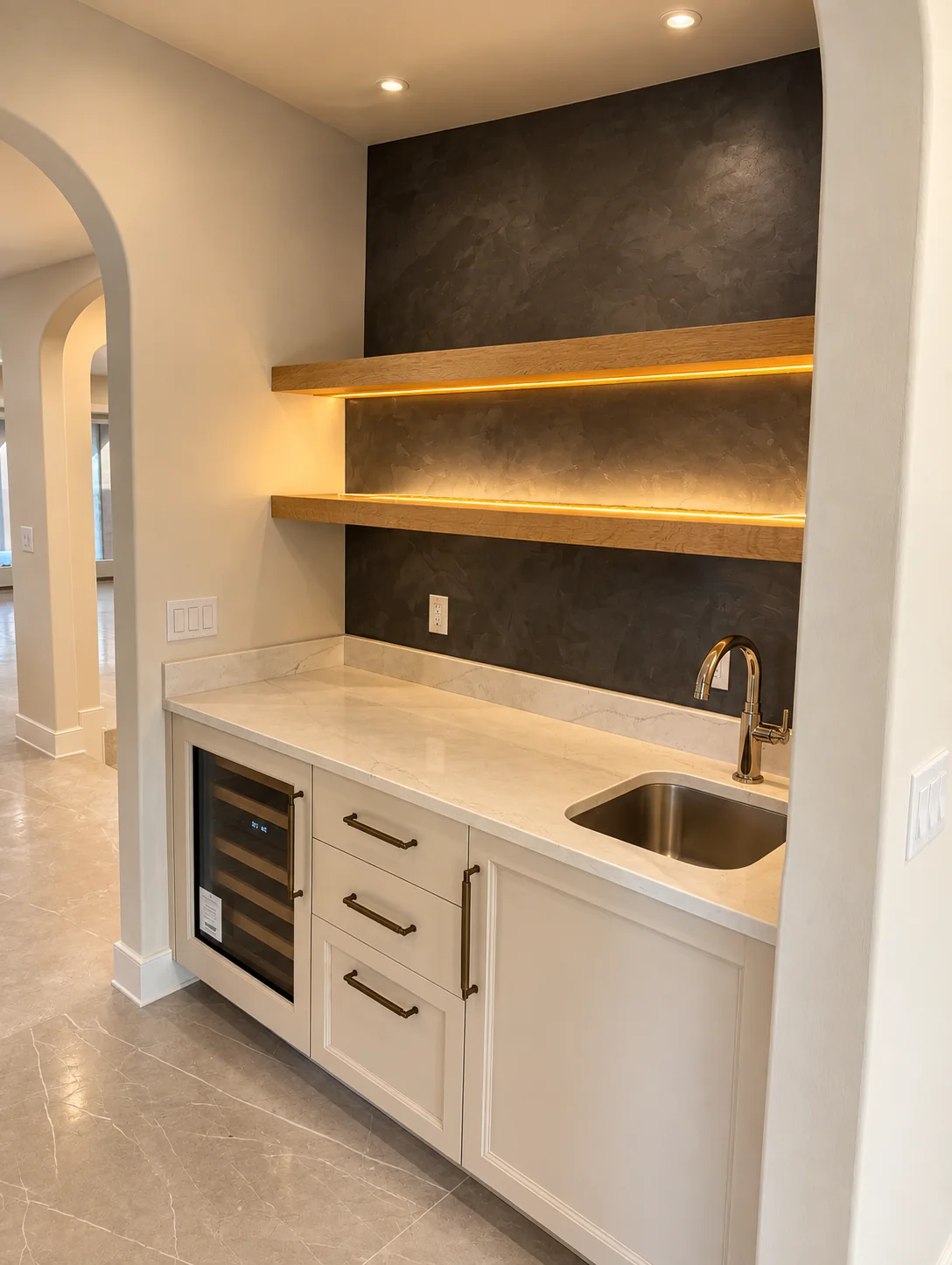 Dark charcoal plaster feature wall behind floating oak shelves with LED lighting above a wet bar with white cabinetry, marble countertop, wine refrigerator and brass faucet in Wayne, NJ